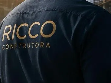 Profissional da RICCO de uniforme e capacete, com o projeto sob o braço, em visita a canteiro.