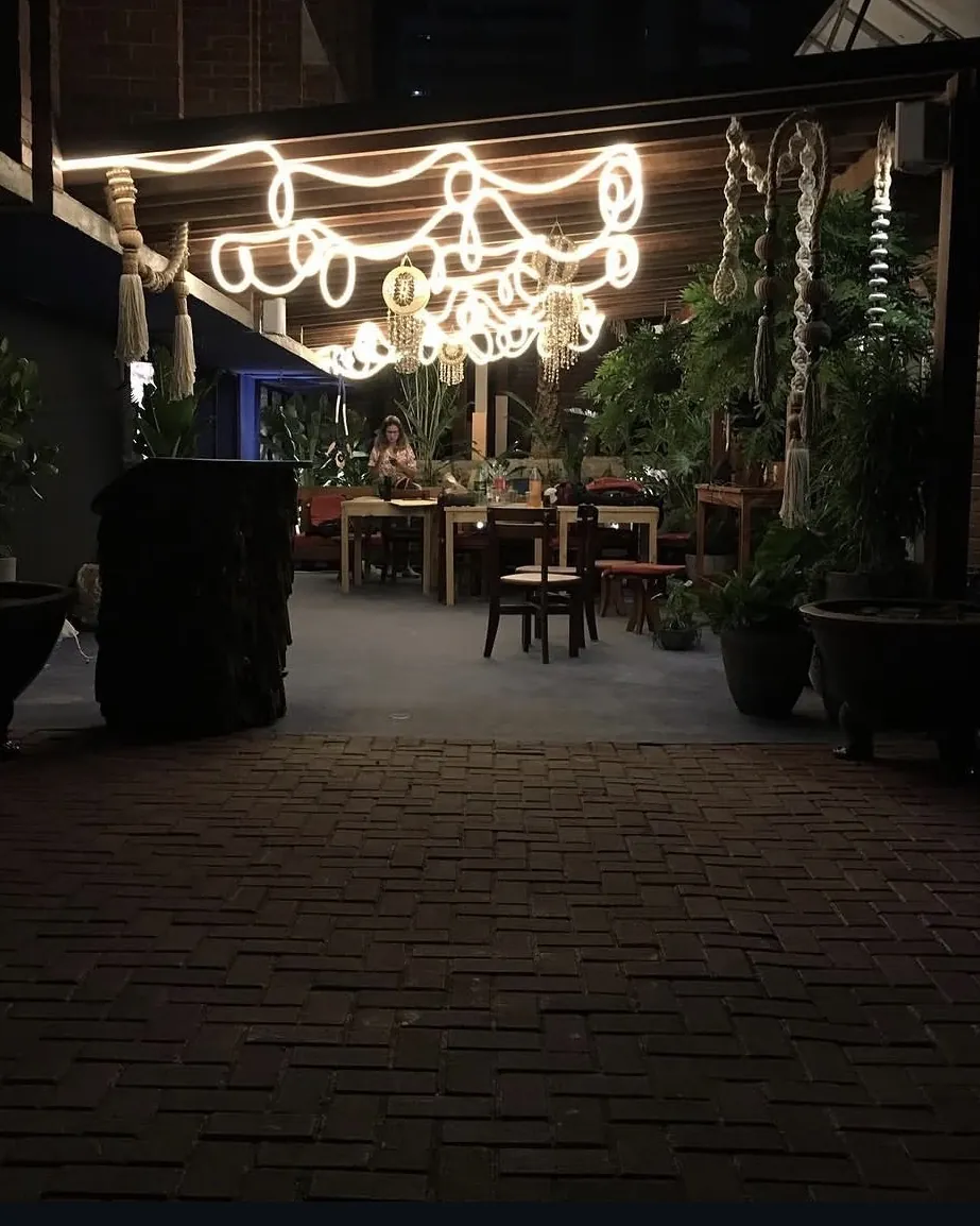 Área externa de restaurante com piso em tijolo aparente, luminárias decorativas suspensas e paisagismo.